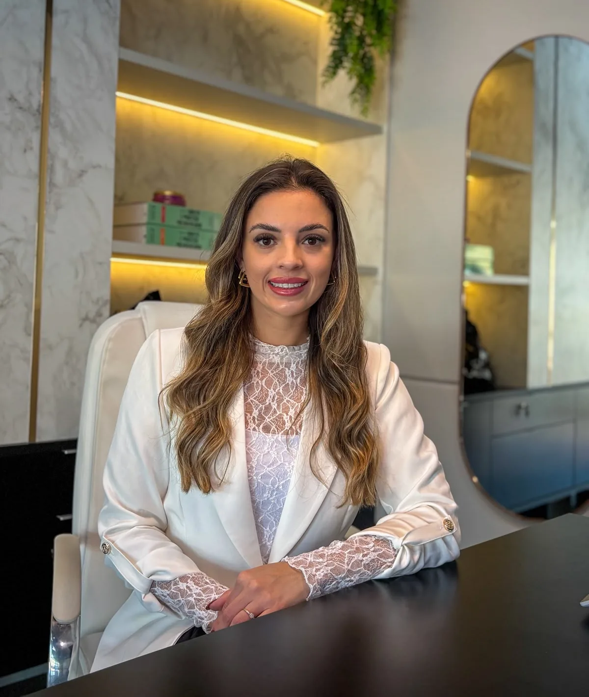 Rafaela Koling sorrindo, sentada à mesa do escritório, de blazer branco.