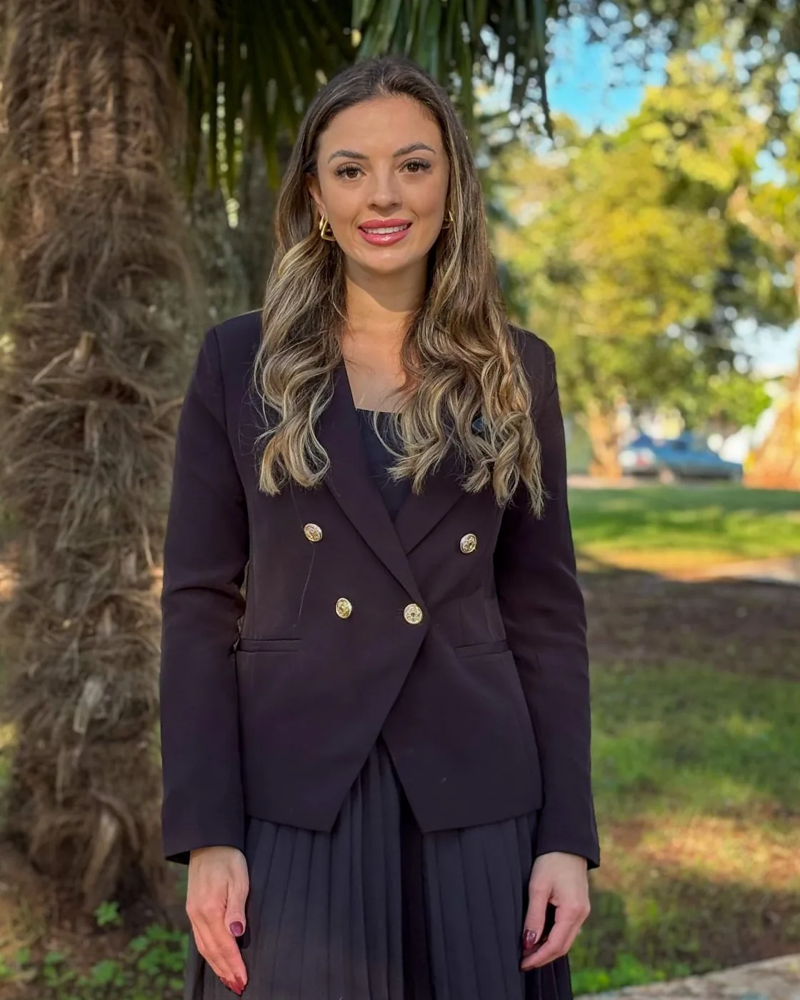 Rafaela Koling de blazer escuro, em pé numa praça arborizada, sorrindo.